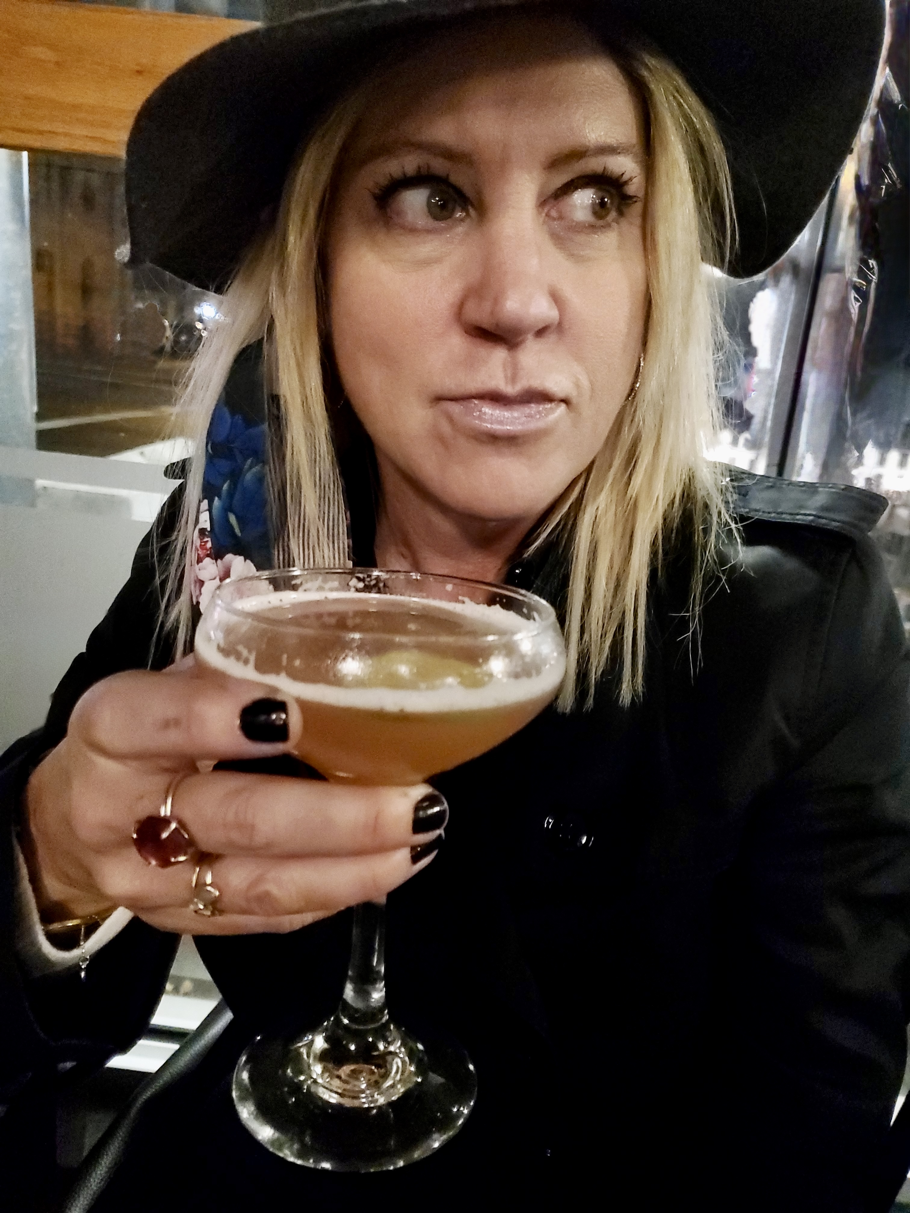 Blond woman with a black hat and a black coat drinking a martini looking to the side.